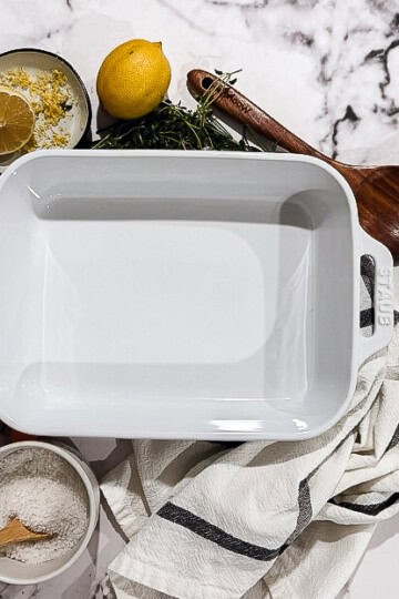 Overhead view of white rectangular baking dish surrounded by fresh herbs, garlic cloves, citrus slices, and autumn botanicals on light marble surface