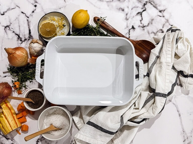 Overhead view of white rectangular baking dish surrounded by fresh herbs, garlic cloves, citrus slices, and autumn botanicals on light marble surface