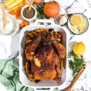Overhead view of golden roasted chicken in white baking dish surrounded by fresh oranges, herbs, and seasonal garnishes on marble surface