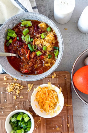 Overhead view of hearty chili in white bowl with colorful toppings including shredded cheese, jalapeños, and fresh garnishes on light surface