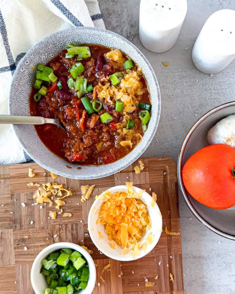 Overhead view of hearty chili in white bowl with colorful toppings including shredded cheese, jalapeños, and fresh garnishes on light surface