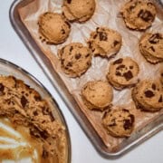 Alt Text: Chocolate chip cookie dough balls on parchment-lined baking sheet next to mixing bowl with wooden spoon